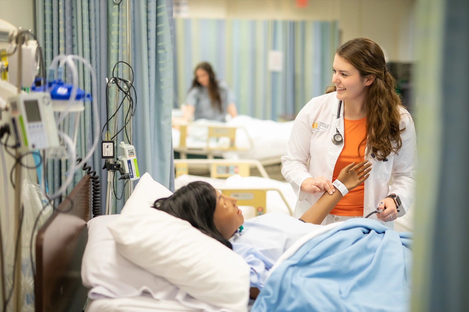 A nurse takes a patient's pulse while smiling and looking at medical readings in a hospital