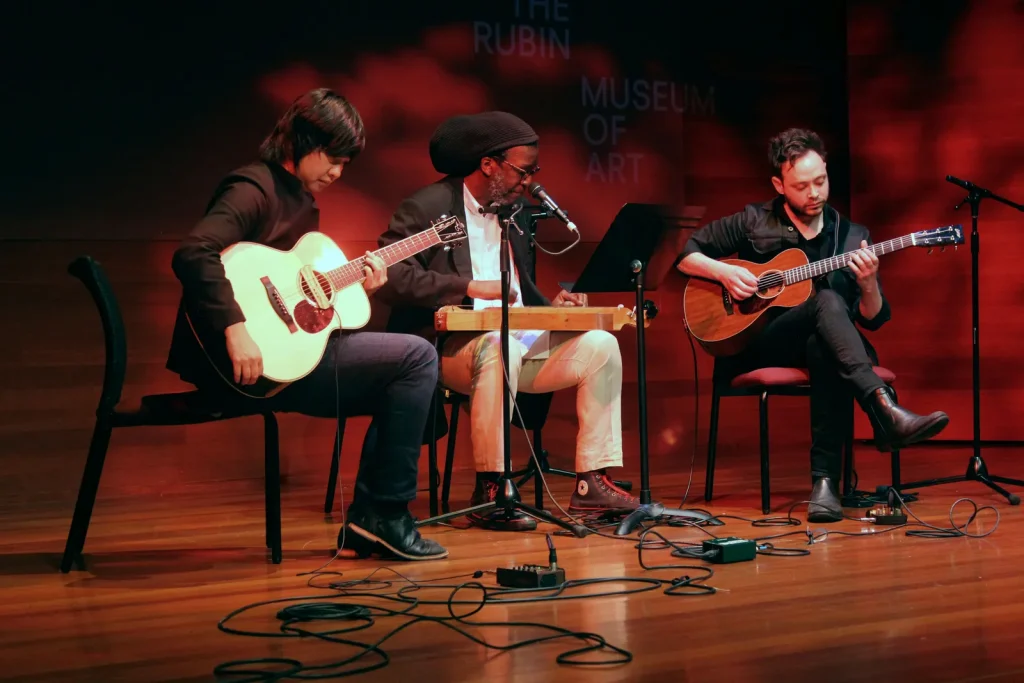 Three musicians on stage, two with guitars and one with a wooden instrument on his lap