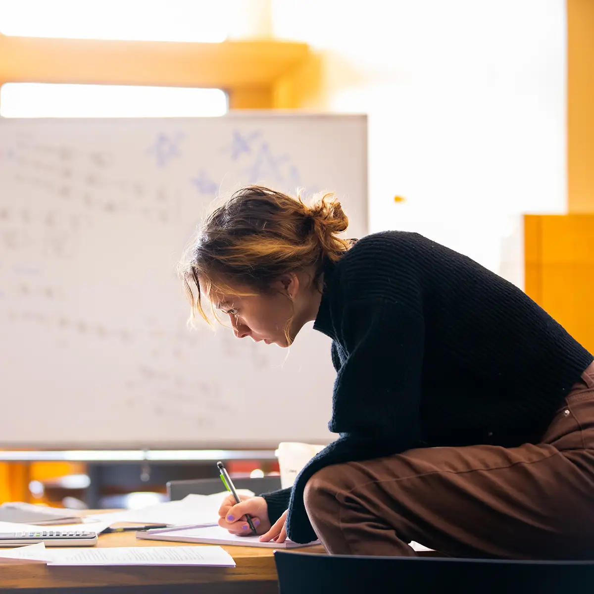 A female student sitting cross legged and working on a project with a white board behind her