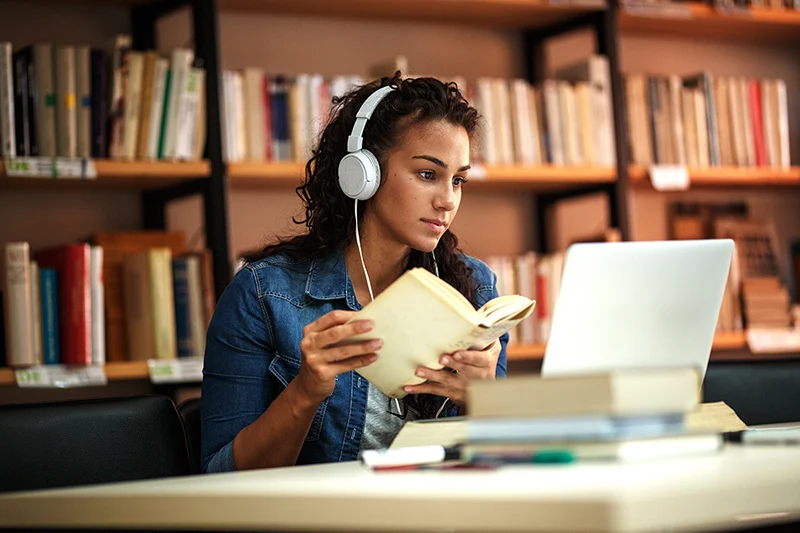 A woman wearing headphones and holding a book while looking at a computer screen in the library