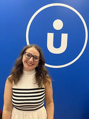 A headshot photo of a young woman in a white dress and wearing glasses with a blue wall and a white logo behind her