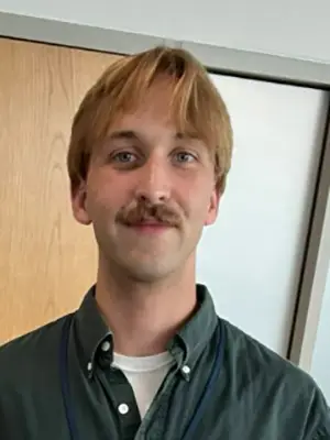 Headshot photo of a young man with a moustache and wearing a green shirt