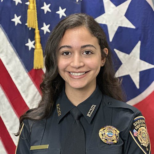A photo of a woman in a county police uniform with the American flag behind her