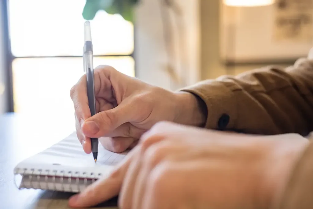 A hand holds a pen that writes against a spiral notebook.