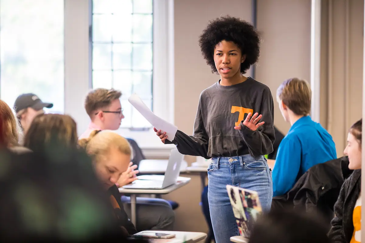 A graduate student stands in a classroom among undergraduate students. 