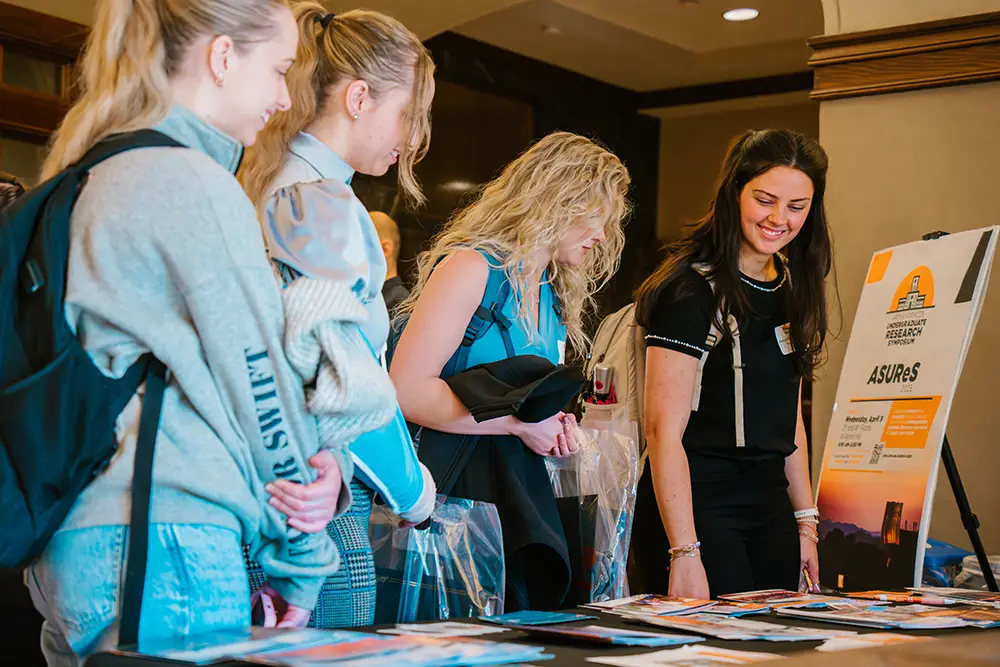 Four students stand at a registration table for the Arts and Sciences Undergraduate Research Symposium (ASUReS) event.