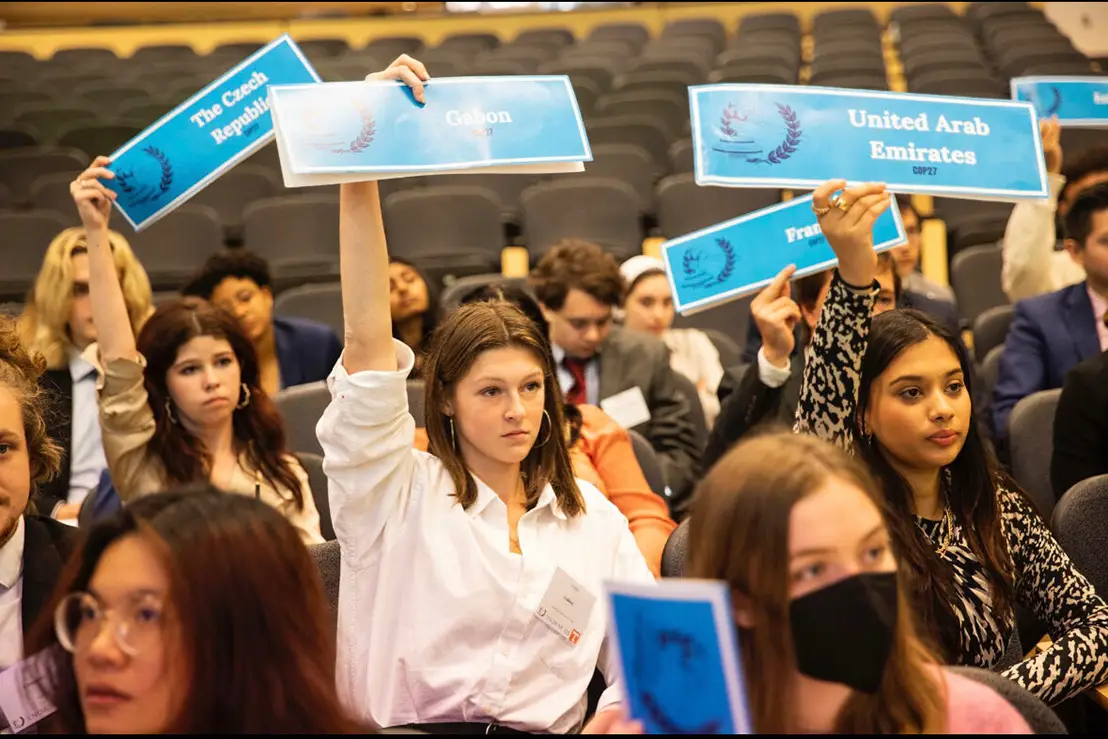 Emma Miller holds a sign that represents the nation of "Gabon" during the Model UN conference, alongside an auditorium filled with other students. 