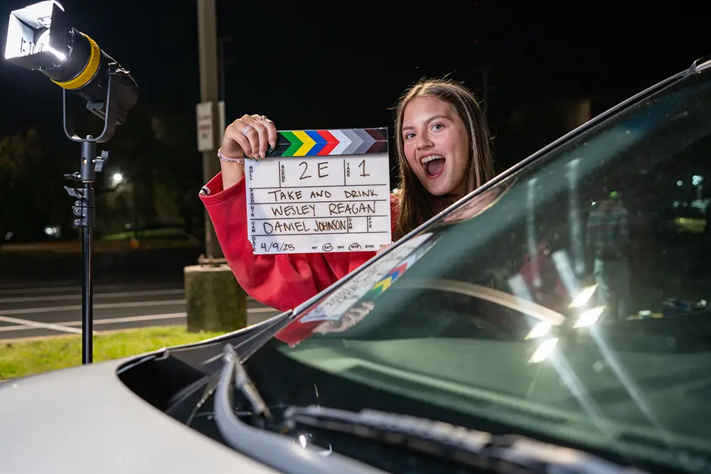 A student filmmaker holds a clapperboard behind a vehicle and lighting equipment
