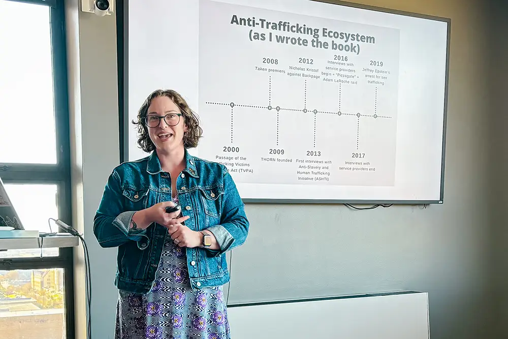 Corinne Schwarz stands next to a project with PowerPoints slides that read, "Anti-Trafficking Ecosystem (as I wrote the book."
