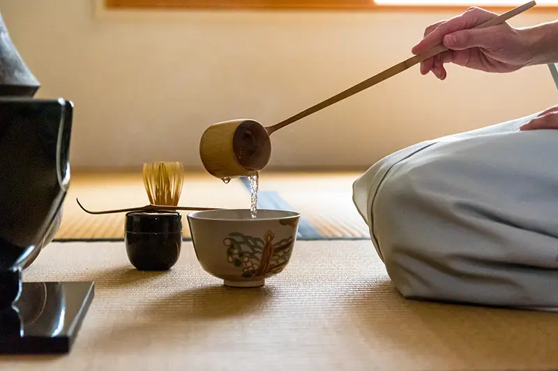 A Japanese woman and tea master uses a Hishaku bamboo ladle to pour hot water into chawan tea bowl. Adobe Stock.