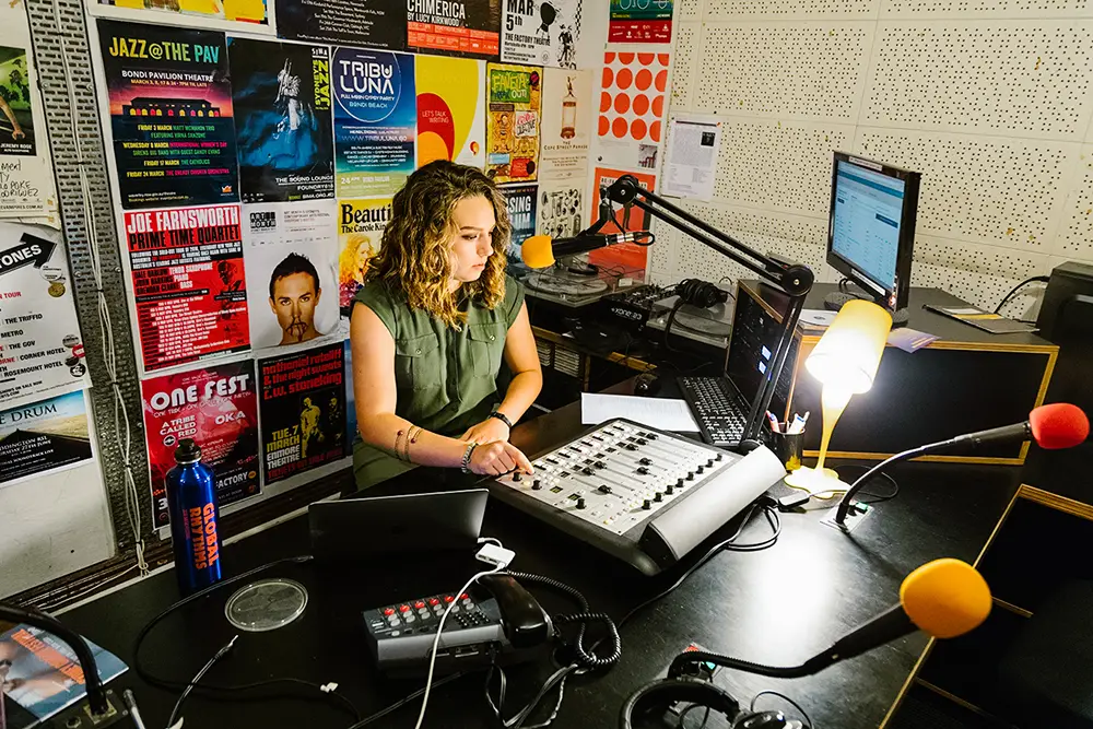 A woman sits at a computer and touches the controls on a soundboard.