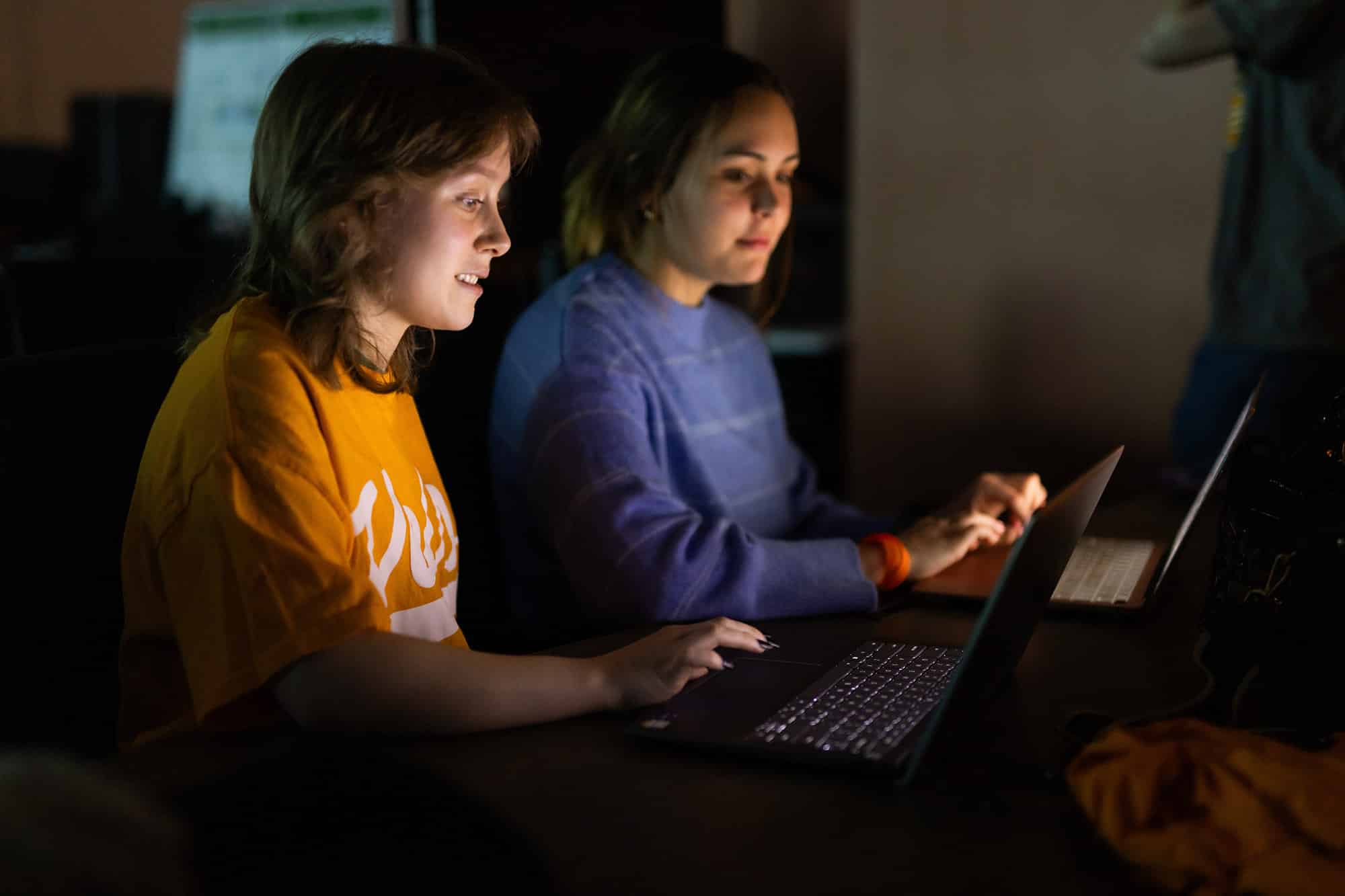 Two students sit in a dark room working on their laptops. 