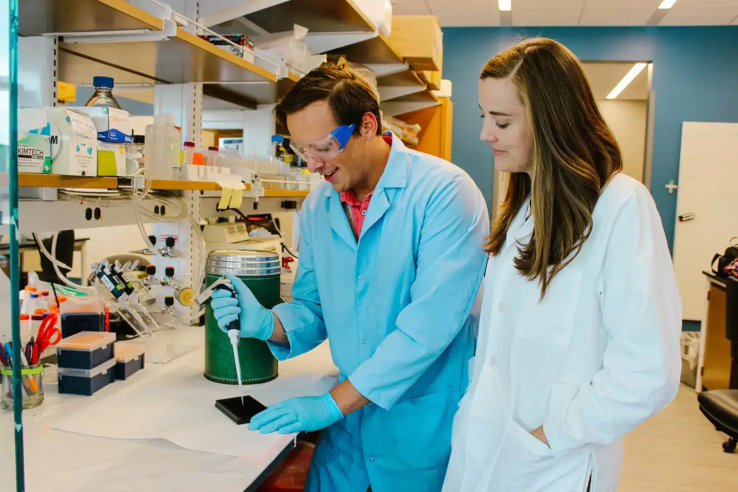 Two students work inside of the lab, placing samples into a try while wearing lab coats and protective glasses. 